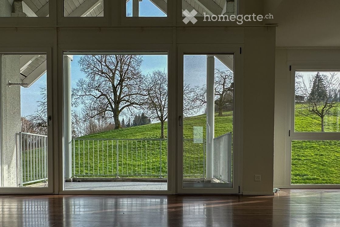 Bright, empty room with large glass doors leading to a balcony overlooking a green, tree-lined hillside. The interior features a polished wooden floor and white walls.