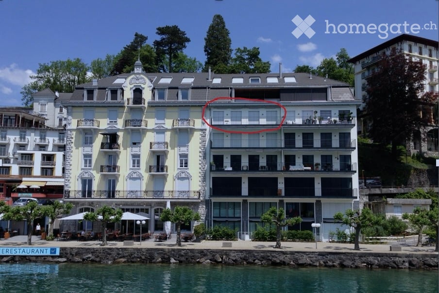 Multi-story building with balconies overlooking a waterfront, surrounded by trees and a restaurant area.