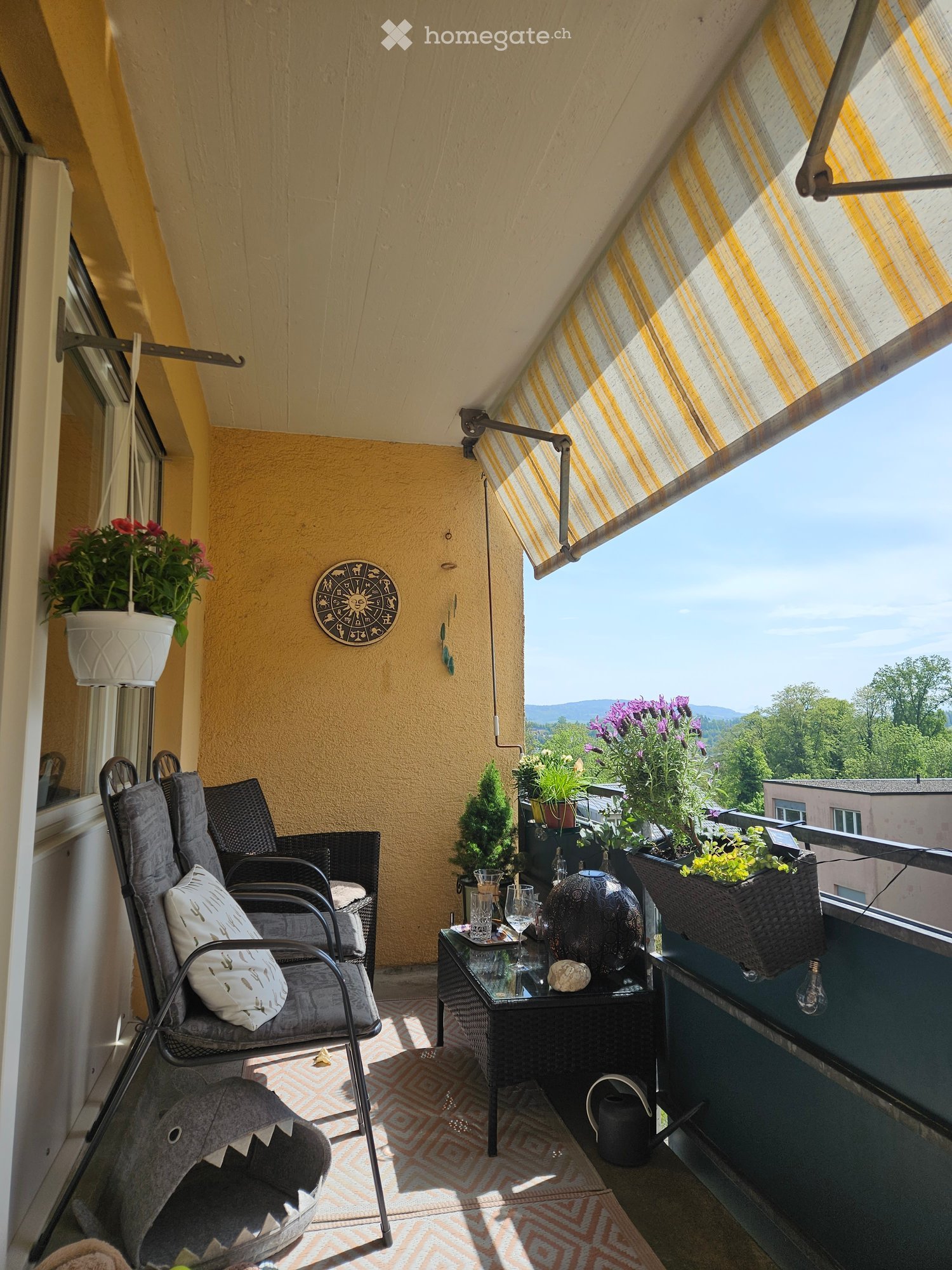 Balcony with yellow walls, grey chairs, small table, potted plants, and a scenic view of greenery and mountains.