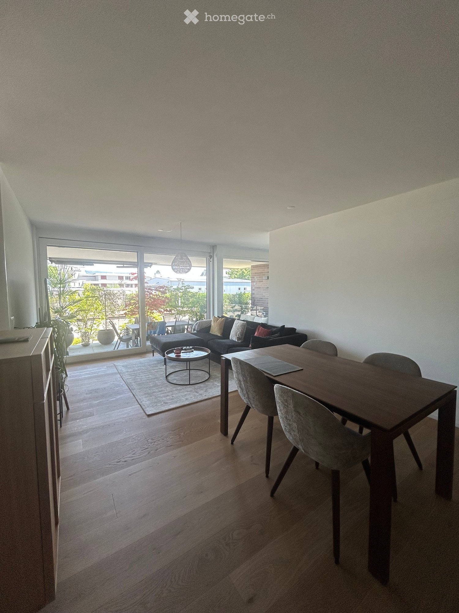Modern living and dining area with wooden flooring, large windows, and a balcony view. Features a sofa, coffee table, dining table, and chairs.
