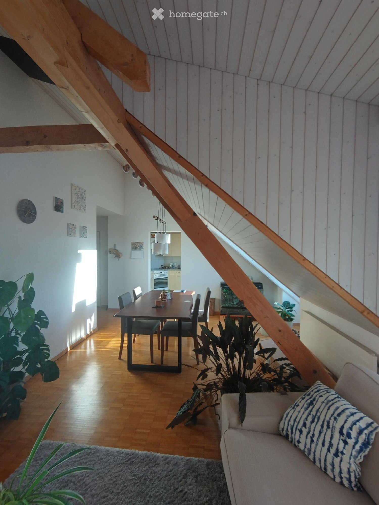 A modern, open-concept living and dining area with wooden floors, a large wooden staircase, a sofa, a dining table with chairs, and several plants.