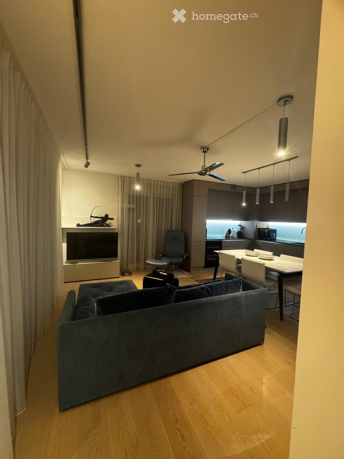 Spacious living room with hardwood floor, gray couch, ceiling fan, large flat screen TV, dining table with chairs, kitchen with black counter, modern design, pendant lighting