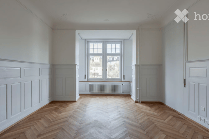 Empty room with wooden herringbone flooring, white walls, large window with radiator below, and white paneling on walls.