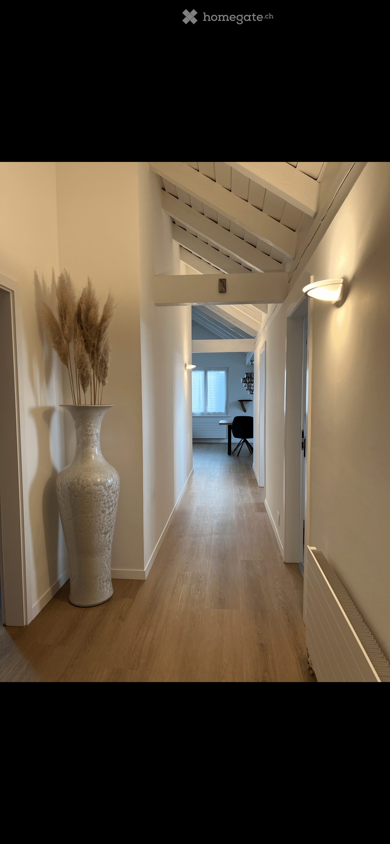 A modern hallway with wooden flooring, white walls, and a large decorative vase on the left side. The hallway leads to a room with a desk and chair visible at the end.