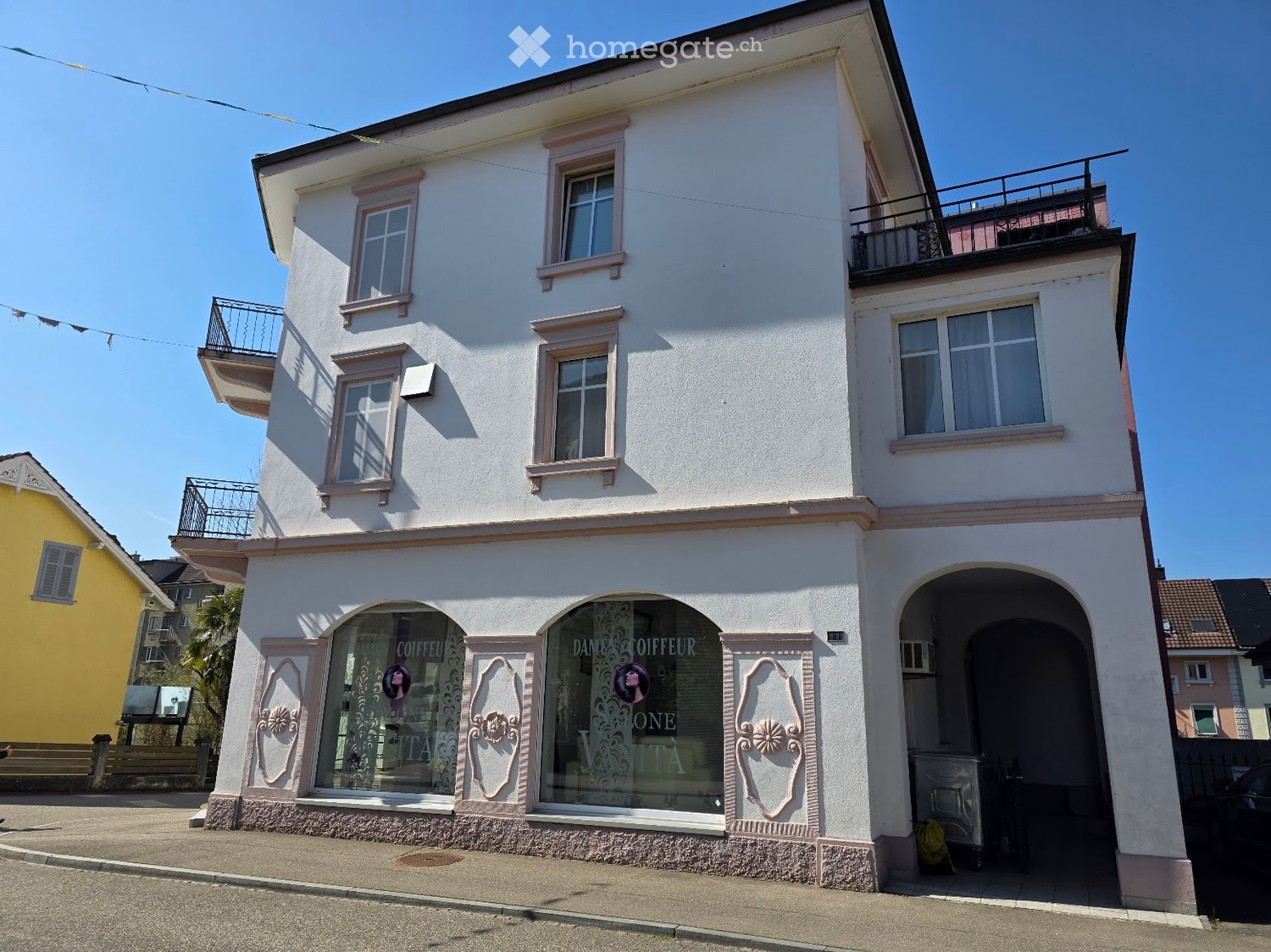 Three-story building with white facade, pink trim, arched storefronts, and balconies on the upper floors.