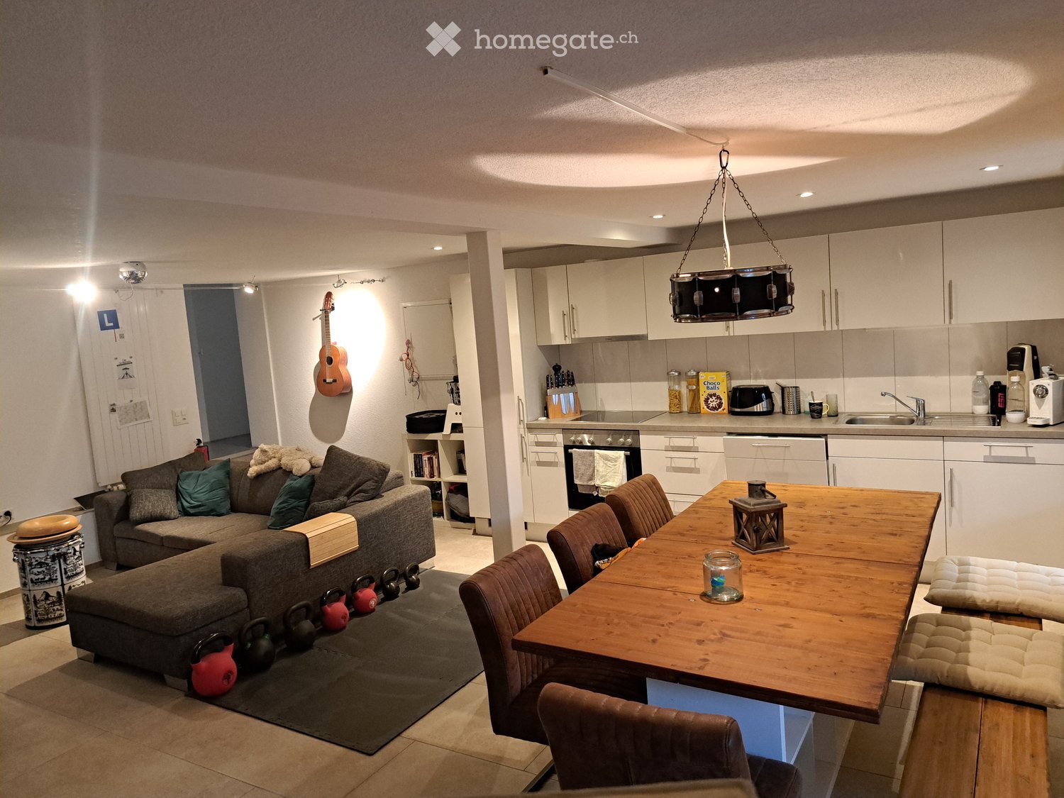 A modern, open-plan living and dining area with a grey sectional sofa, wooden dining table, four brown chairs, a guitar on the wall, and a fully equipped kitchen with white cabinets, stainless steel appliances, and various kitchen items on the counter.