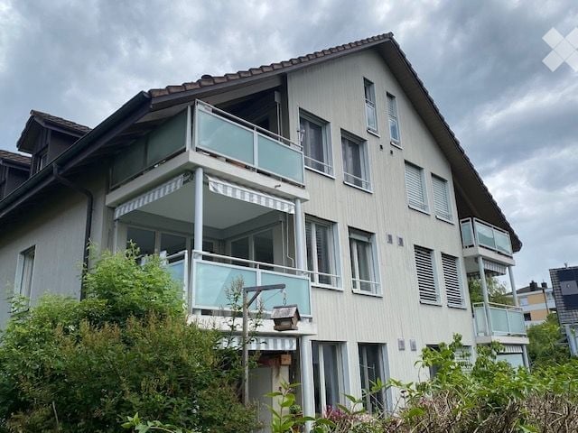 Modern two-story house with light-colored siding, multiple windows, and balconies with glass railings.