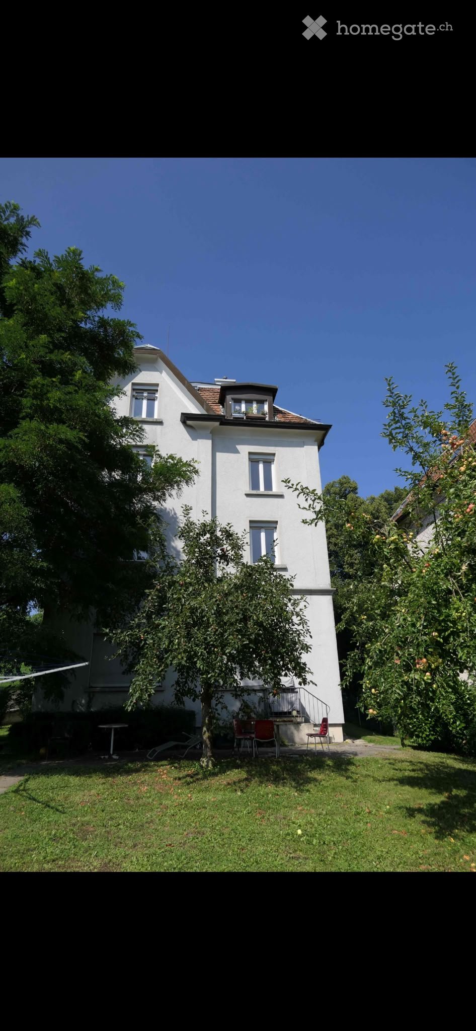 A two-story white house with a pitched roof, surrounded by a well-maintained garden with fruit trees and outdoor seating.