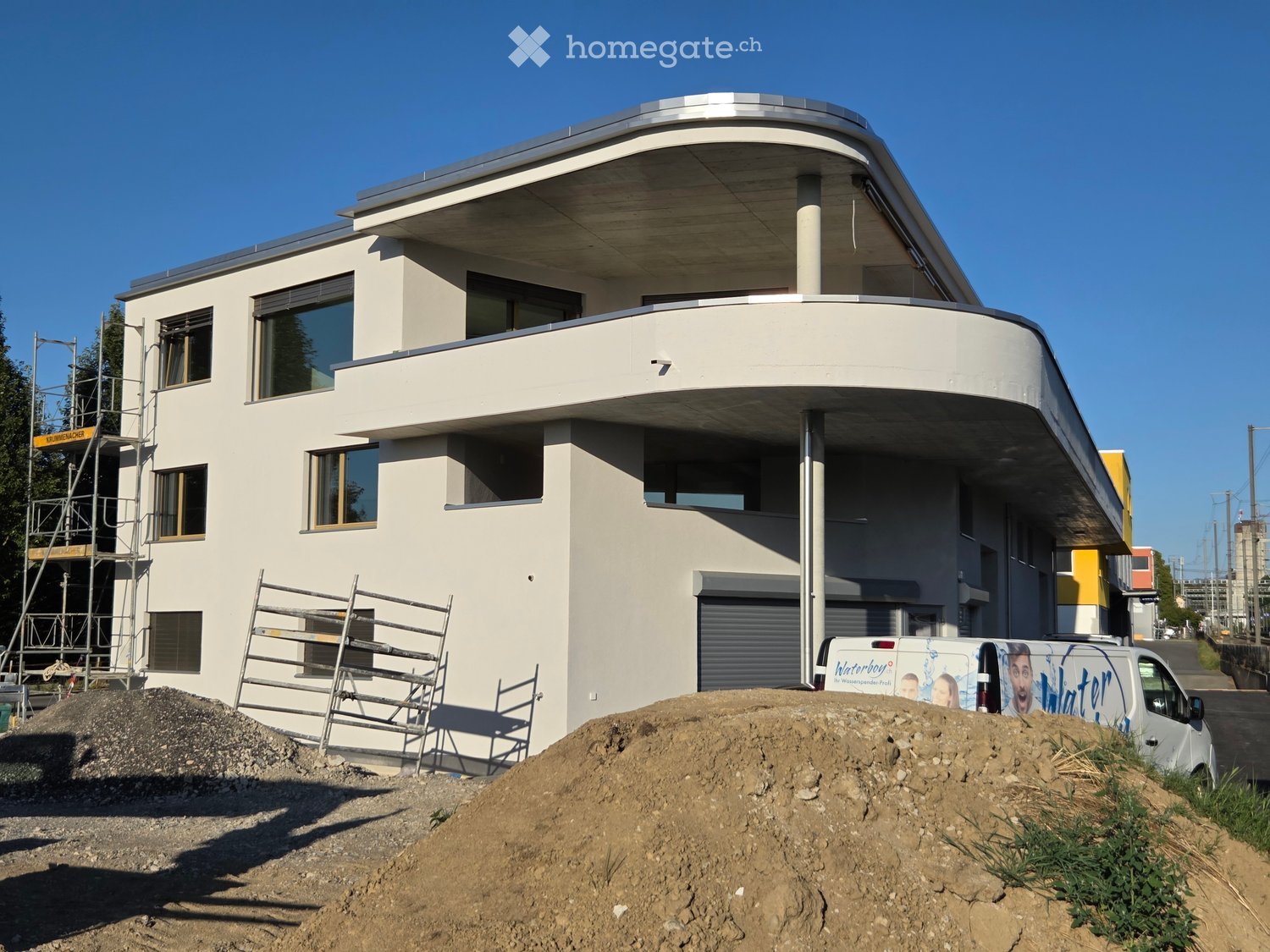 Modern two-story house with white exterior, large windows, and a flat roof. Scaffolding and construction materials are visible, indicating ongoing work.