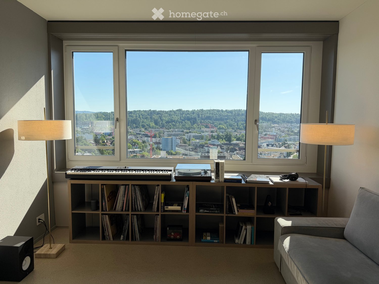 Modern living room with large windows offering a scenic city view, featuring a grey sofa, a wooden bookshelf filled with books and records, a record player, a keyboard, and two floor lamps.