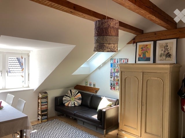 Cozy attic room with slanted wooden ceiling beams, exposed wooden beams, large skylight window, black leather sofa, colorful throw pillow, small dining table with chairs, books stacked on floor, and a tall cabinet with picture frames on top.