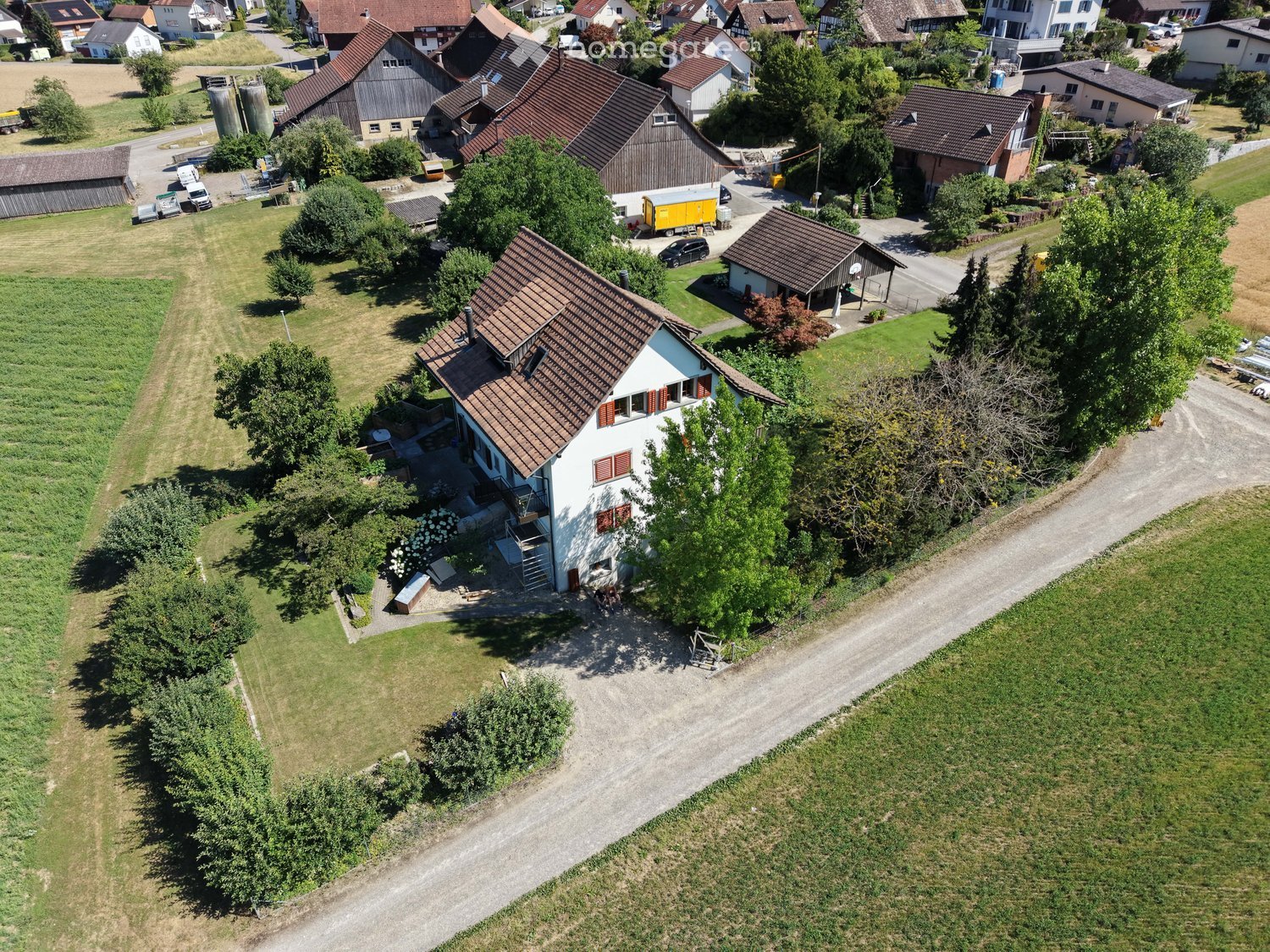Aerial view of a two-story house with a brown tiled roof, surrounded by a well-maintained garden and trees. The house features multiple windows and a small balcony. The surrounding area includes other houses, a road, and open fields.