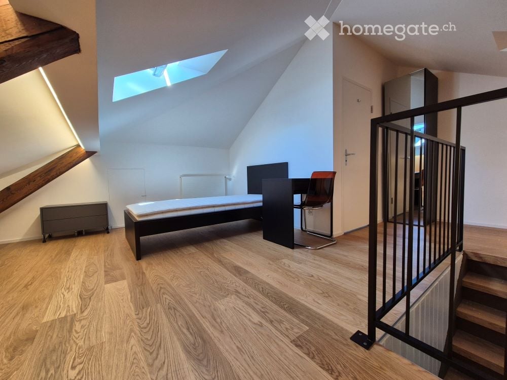 Modern attic bedroom with wooden flooring, slanted ceiling, skylight, bed, chair, table, and wardrobe.