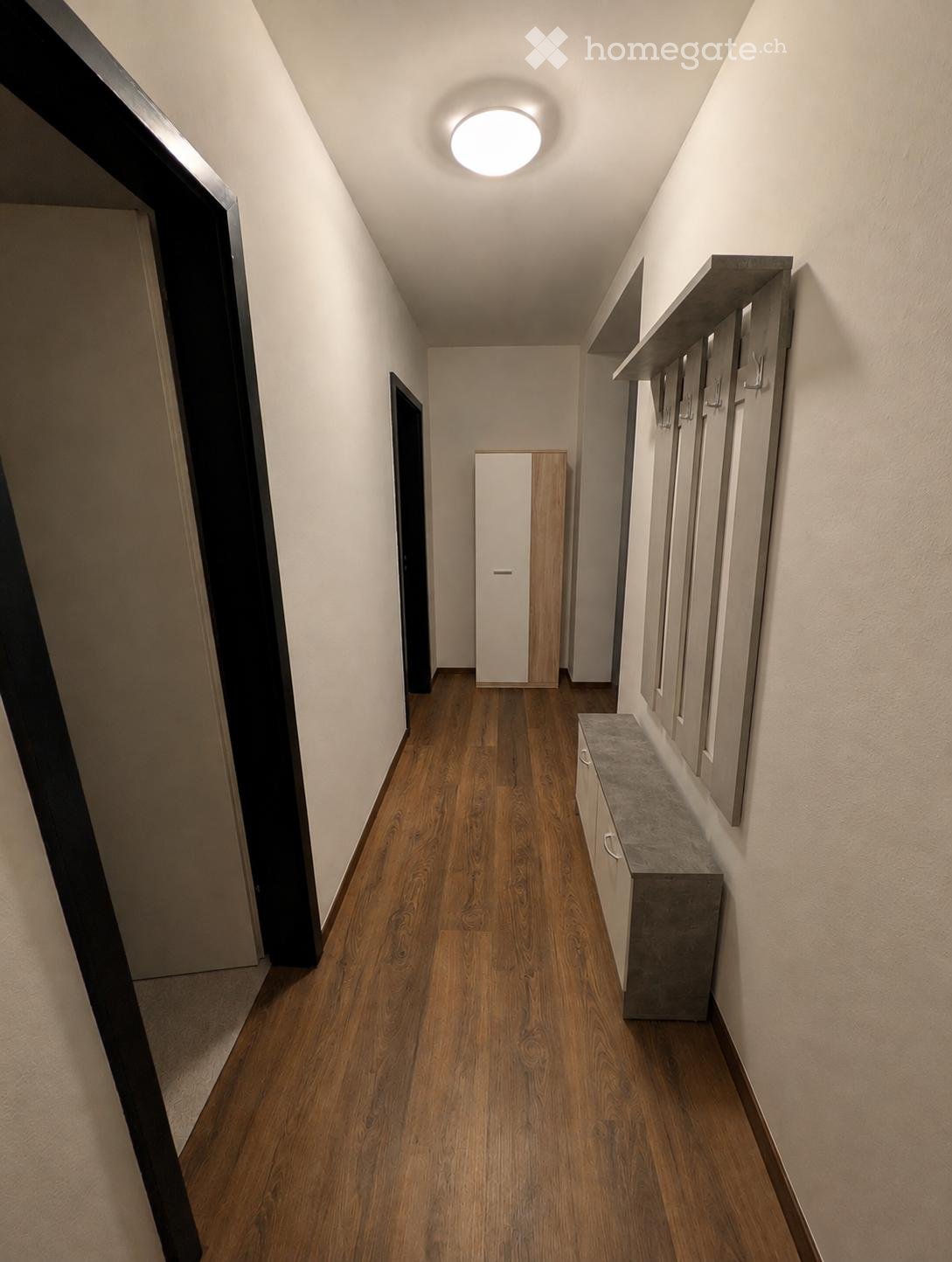 A narrow hallway with wooden flooring, white walls, built-in storage units, and a small bench with a countertop. The ceiling features a round recessed light fixture.