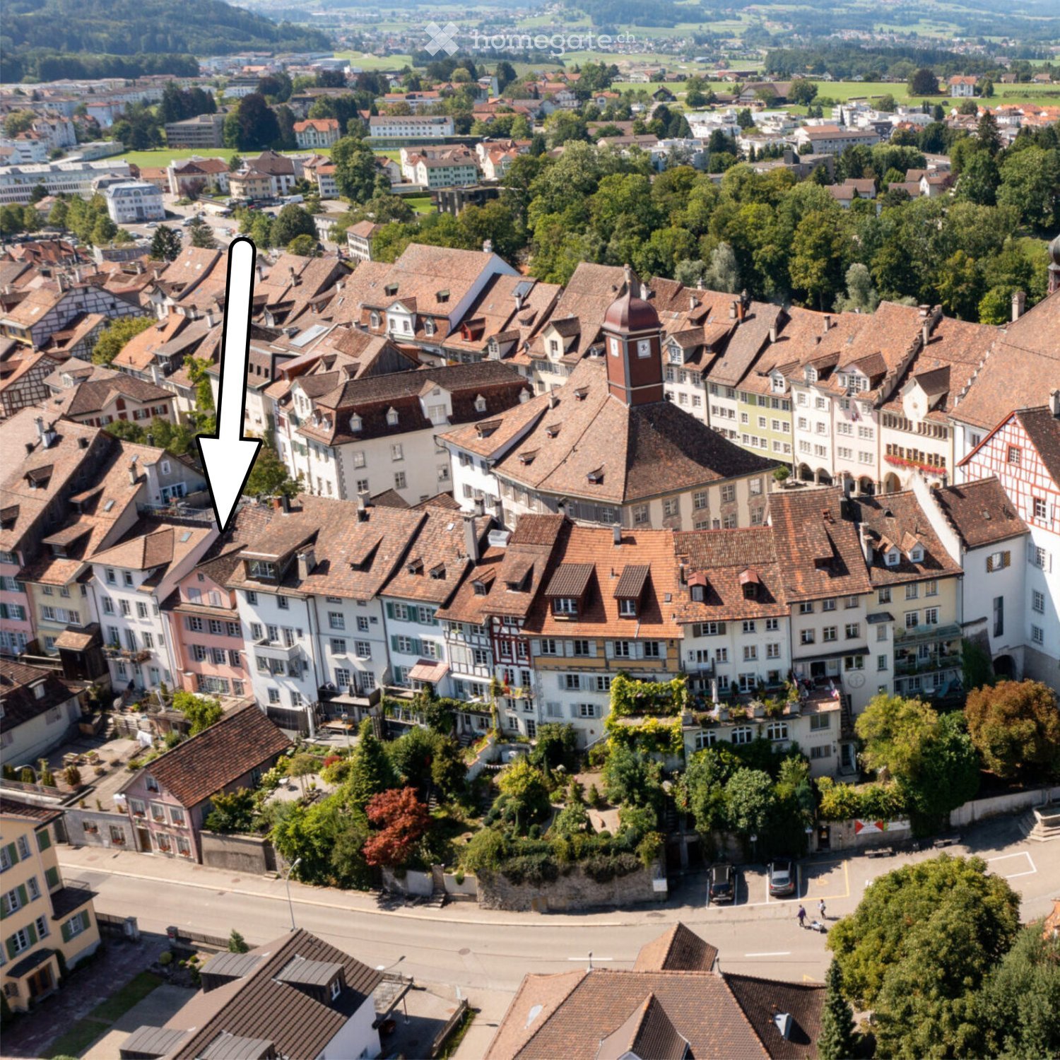 Aerial view of a town with numerous buildings, predominantly featuring red-tiled roofs and white or pastel-colored facades, surrounded by greenery and open fields.