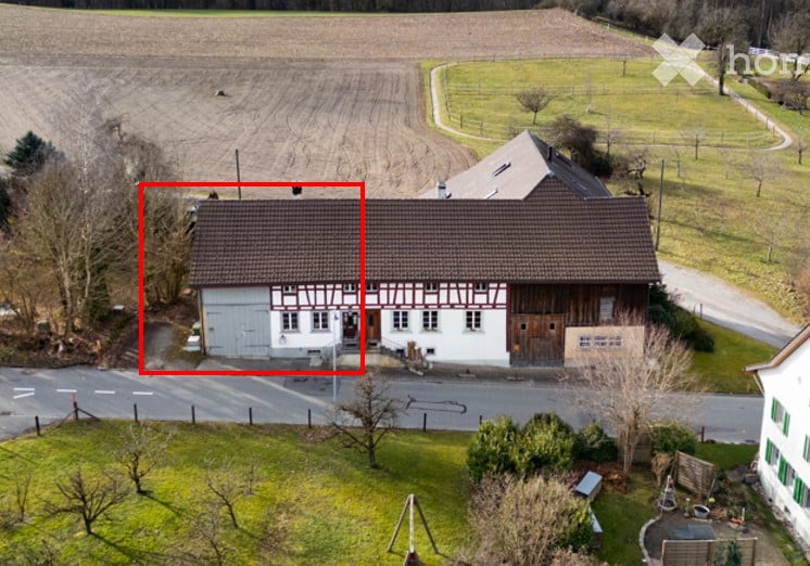 Aerial view of a two-story house with a garage, surrounded by a grassy area and a field in the background.