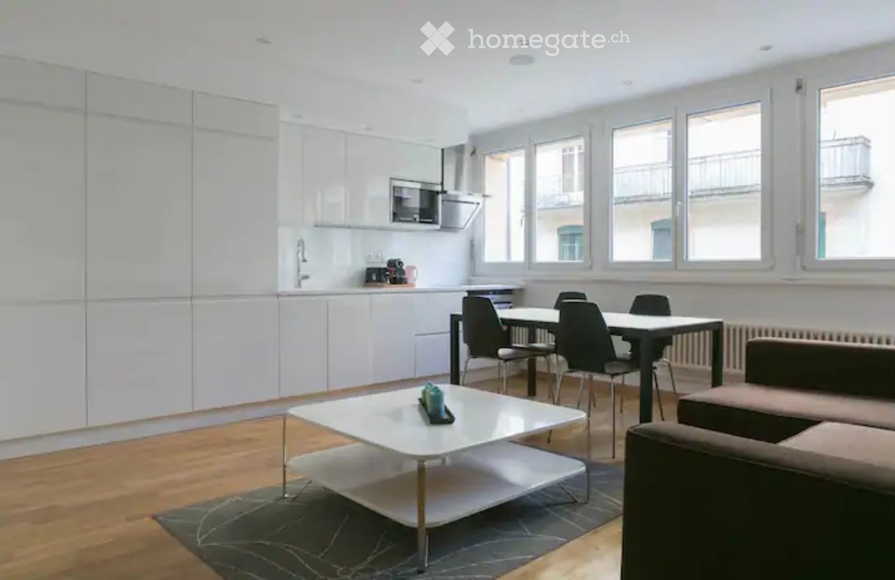 Spacious open-plan living room and kitchen with modern, minimalist design. The kitchen features white cabinets and appliances, including a dishwasher. The living area has a large white coffee table and dining table with black chairs. The room is well-lit