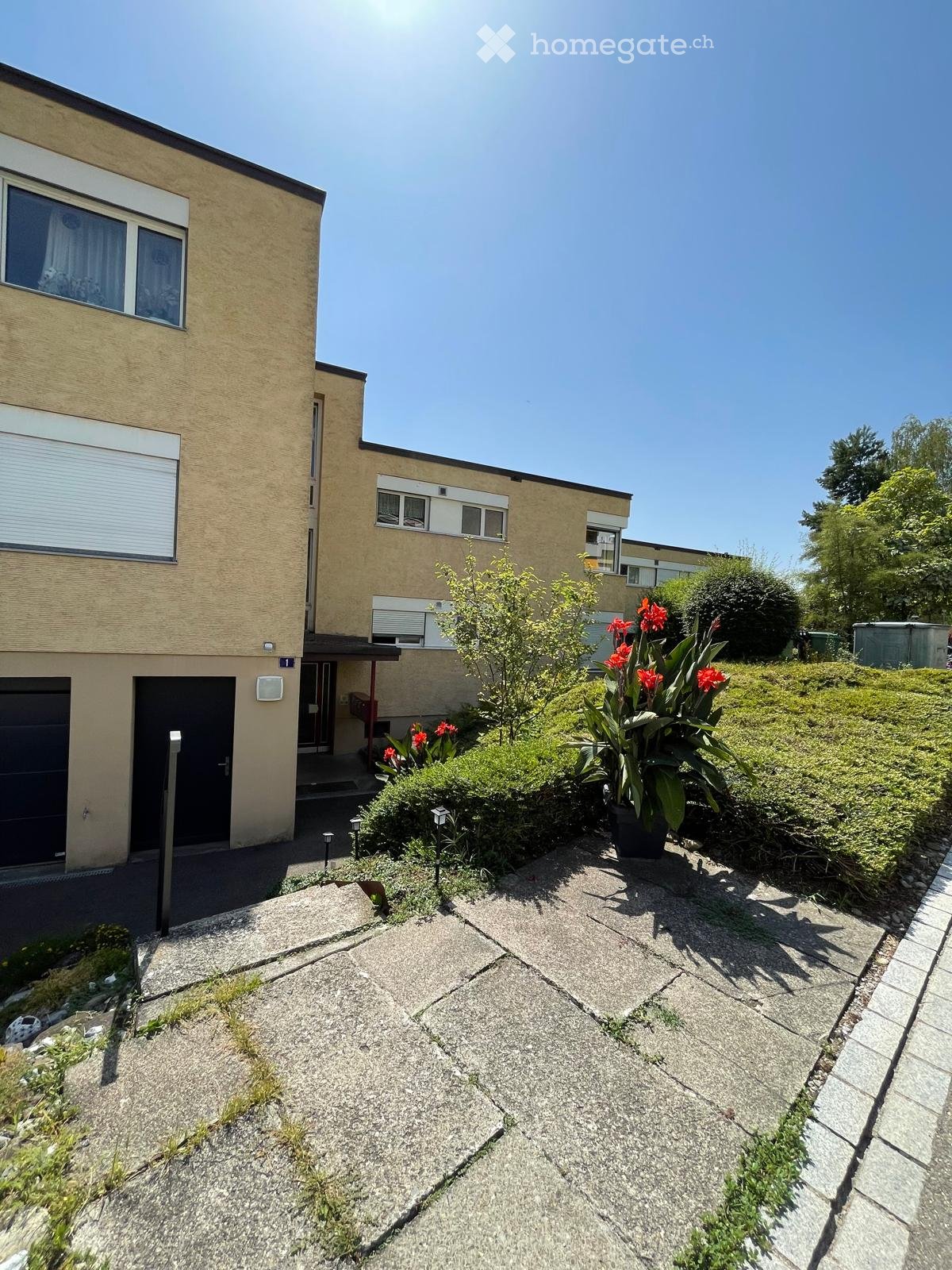Two-story house with beige exterior, garage, windows, and small garden area with plants and flowers.