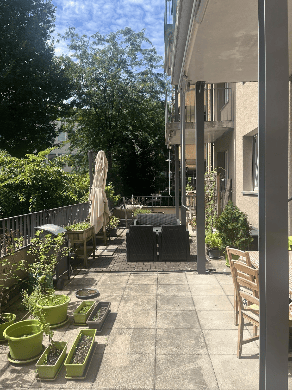Balcony or patio area with potted plants, outdoor furniture, and a grill. The space is surrounded by a metal railing and has a view of greenery.