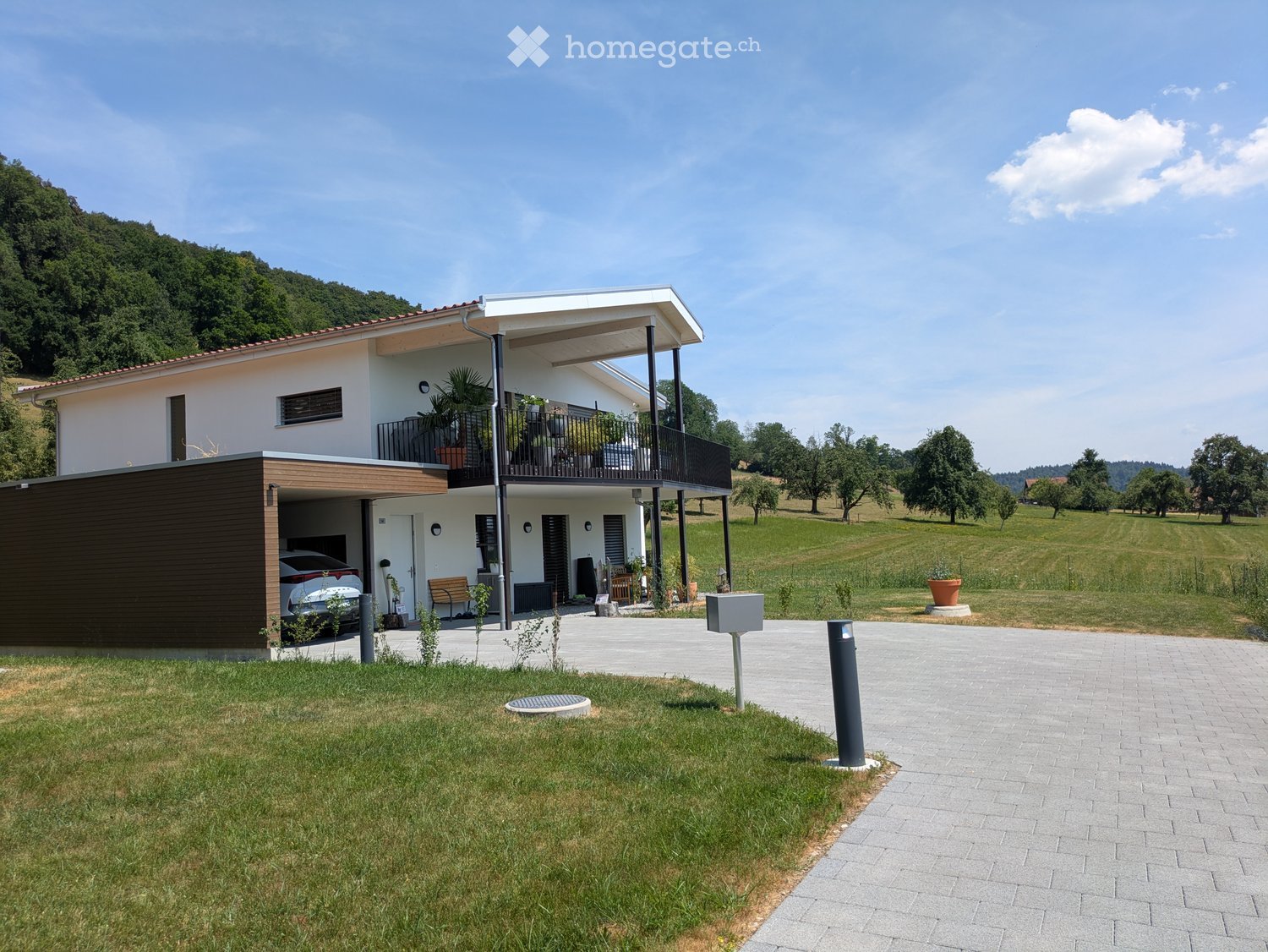 Modern two-story house with white exterior, red-tiled roof, balcony, garage, and surrounding green landscape.