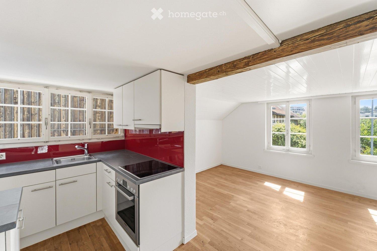 Bright, modern kitchen with white cabinets, stainless steel appliances, and a red backsplash. Features wooden flooring and large windows providing natural light.