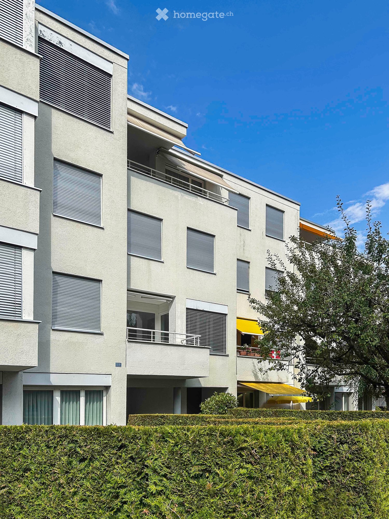 Modern multi-story building with white facade, multiple windows with blinds, balconies, and yellow awnings.