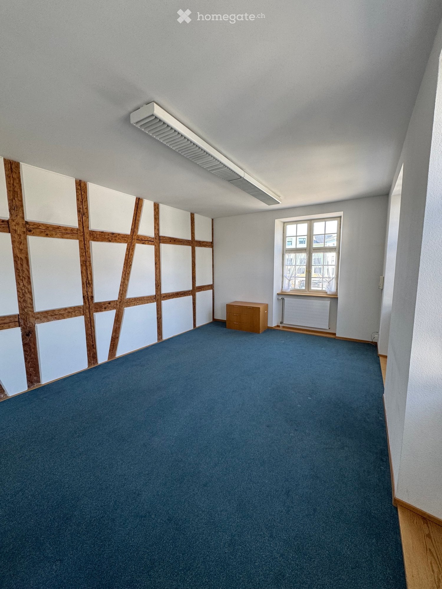 Empty room with blue carpet, white walls, wooden paneling, a window, and a small wooden cabinet.