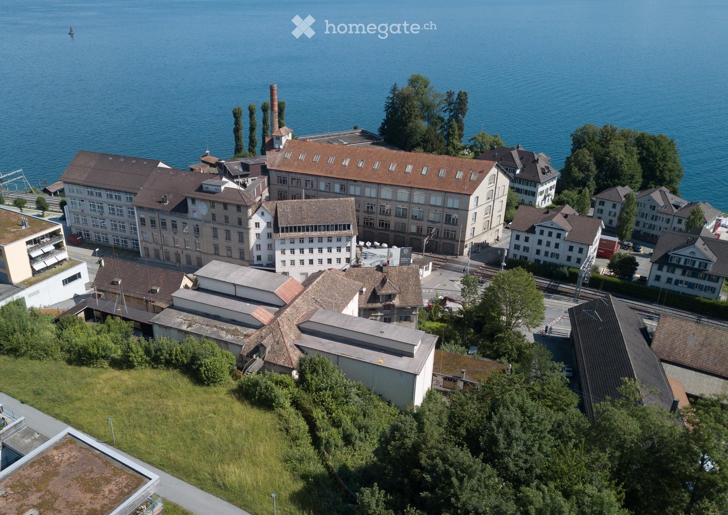 Aerial view of a complex of buildings near a body of water, with multiple structures, chimneys, and surrounding greenery.