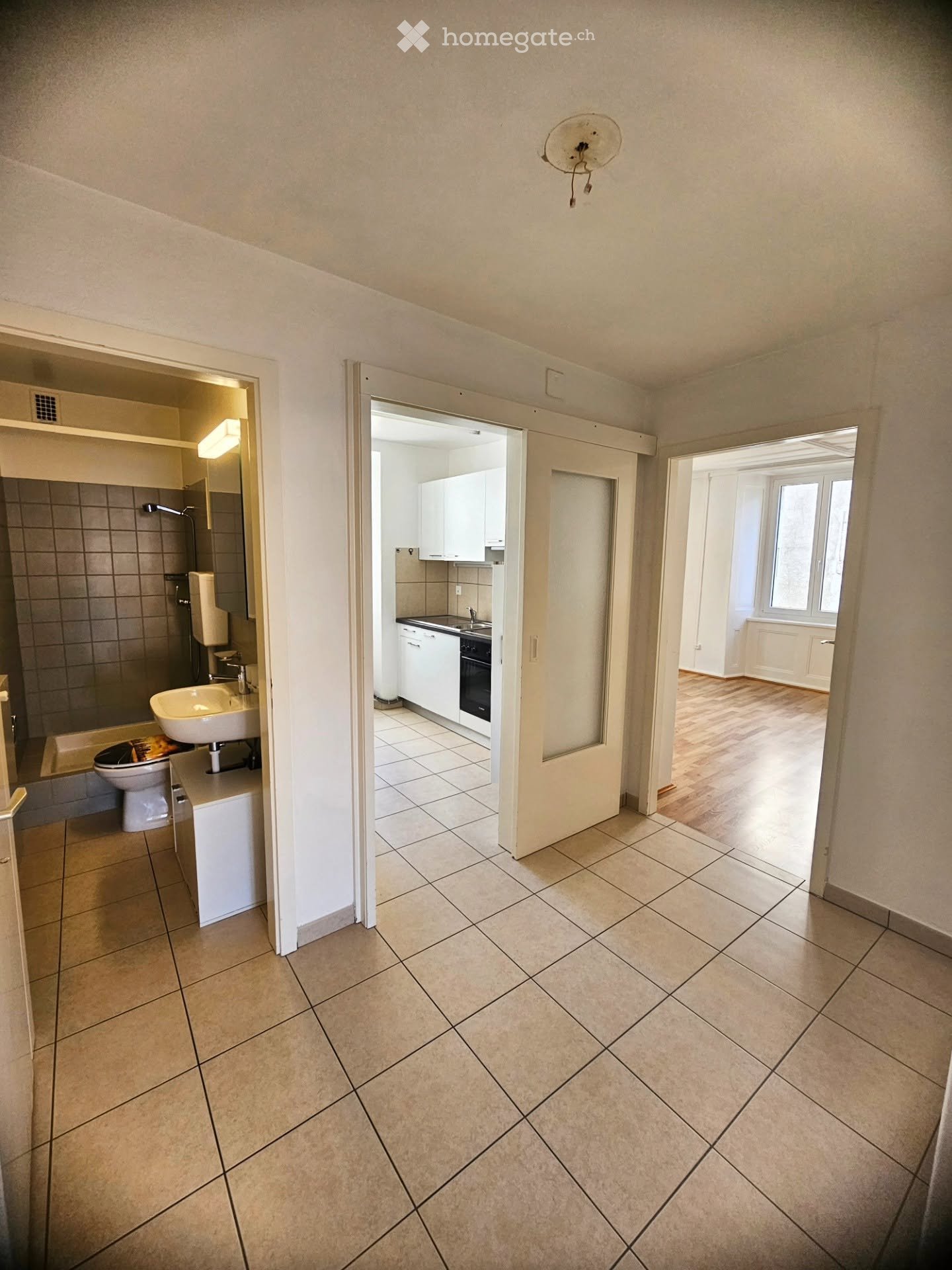 Empty hallway with tiled floor, leading to a bathroom and kitchen area. The bathroom has a toilet, sink, and bathtub, while the kitchen features white cabinets and a sink.