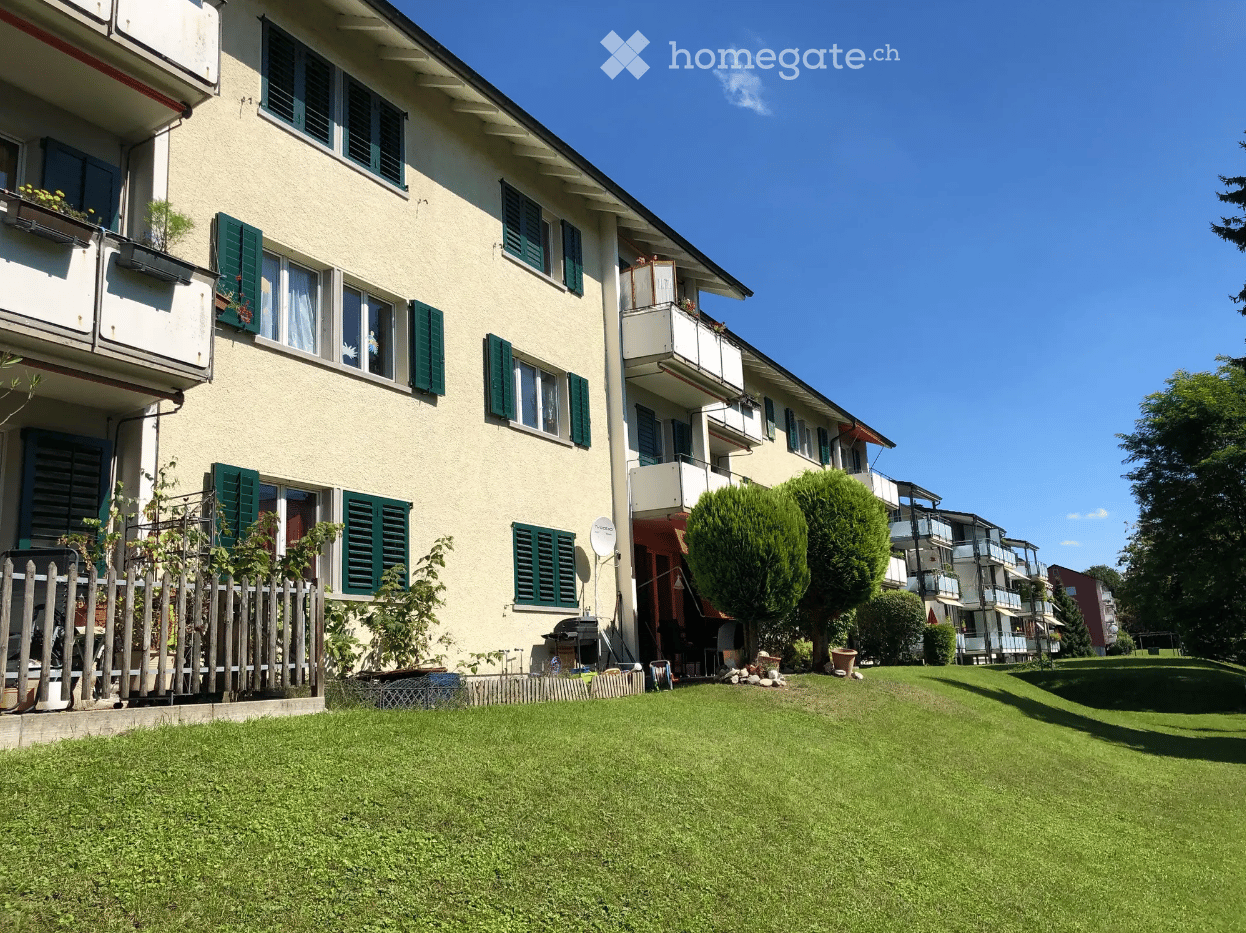 Multi-story residential building with green shutters, balconies, and a well-maintained lawn in front.