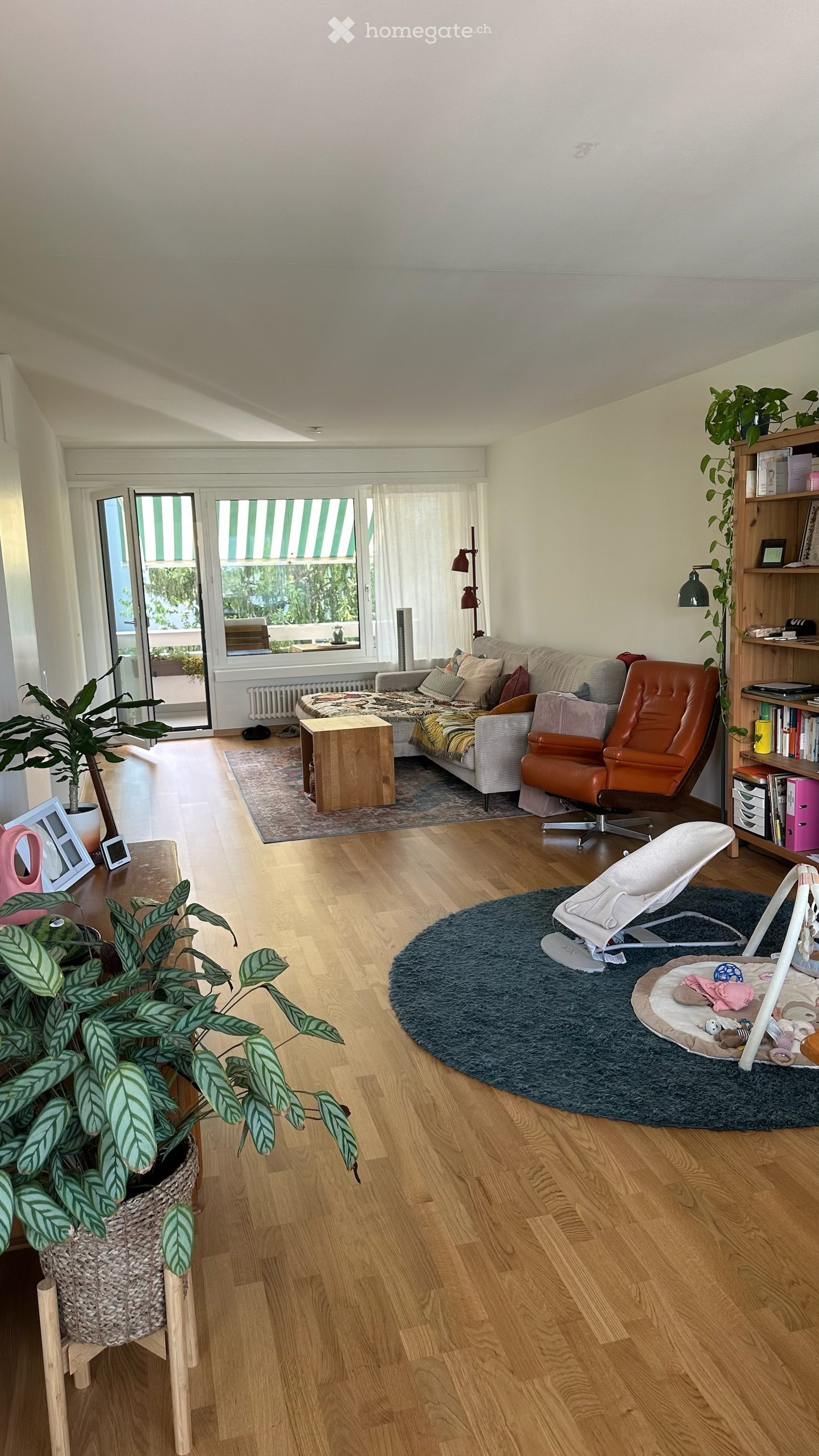 Bright living room with wooden flooring, large windows, and a balcony. Features a sofa, coffee table, armchair, bookshelf, baby chair, and plants.