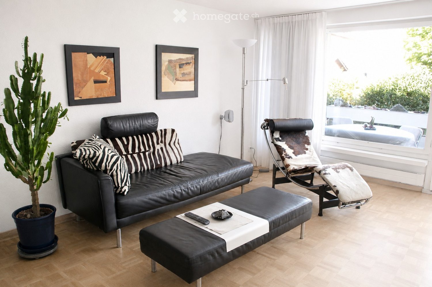 Modern living room with a black leather sofa, zebra-patterned cushions, a white leather stool, a cowhide chair, a potted cactus, and artwork on the walls.