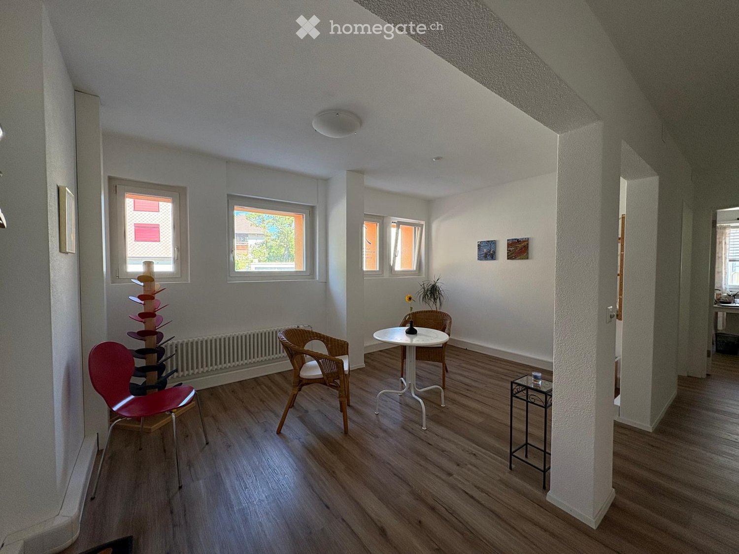 Bright living room with wooden flooring, large windows, a red chair, a white table with a vase, and a small side table.