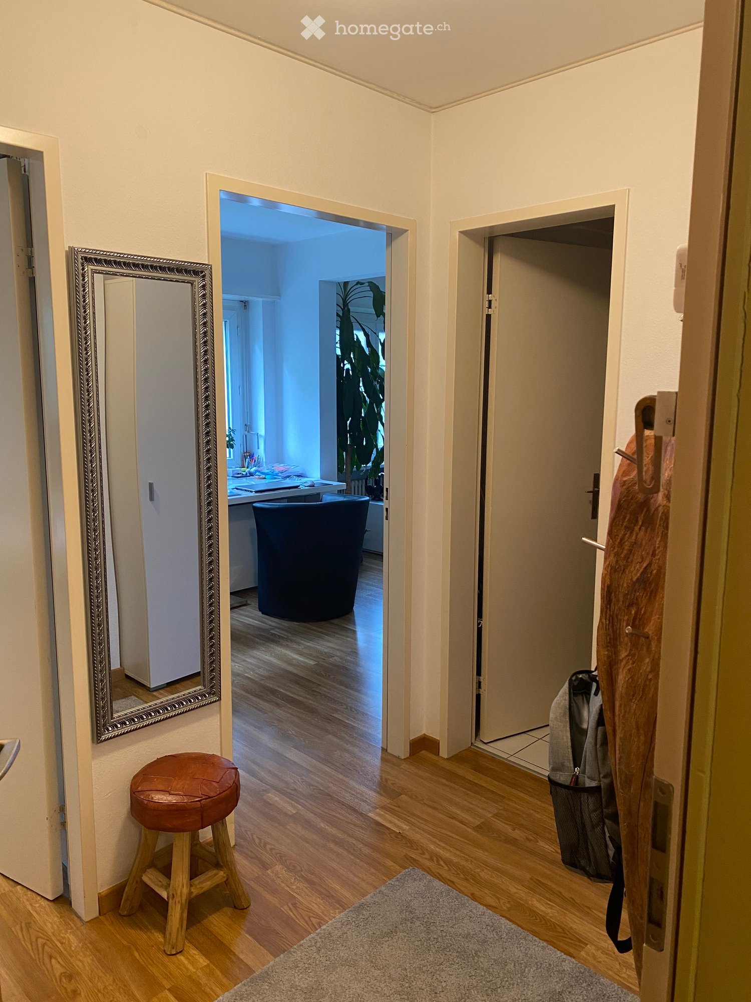 Hallway with wooden flooring, a large mirror on the left wall, a small wooden stool, and an open door leading to a room with a dark blue chair and a desk.