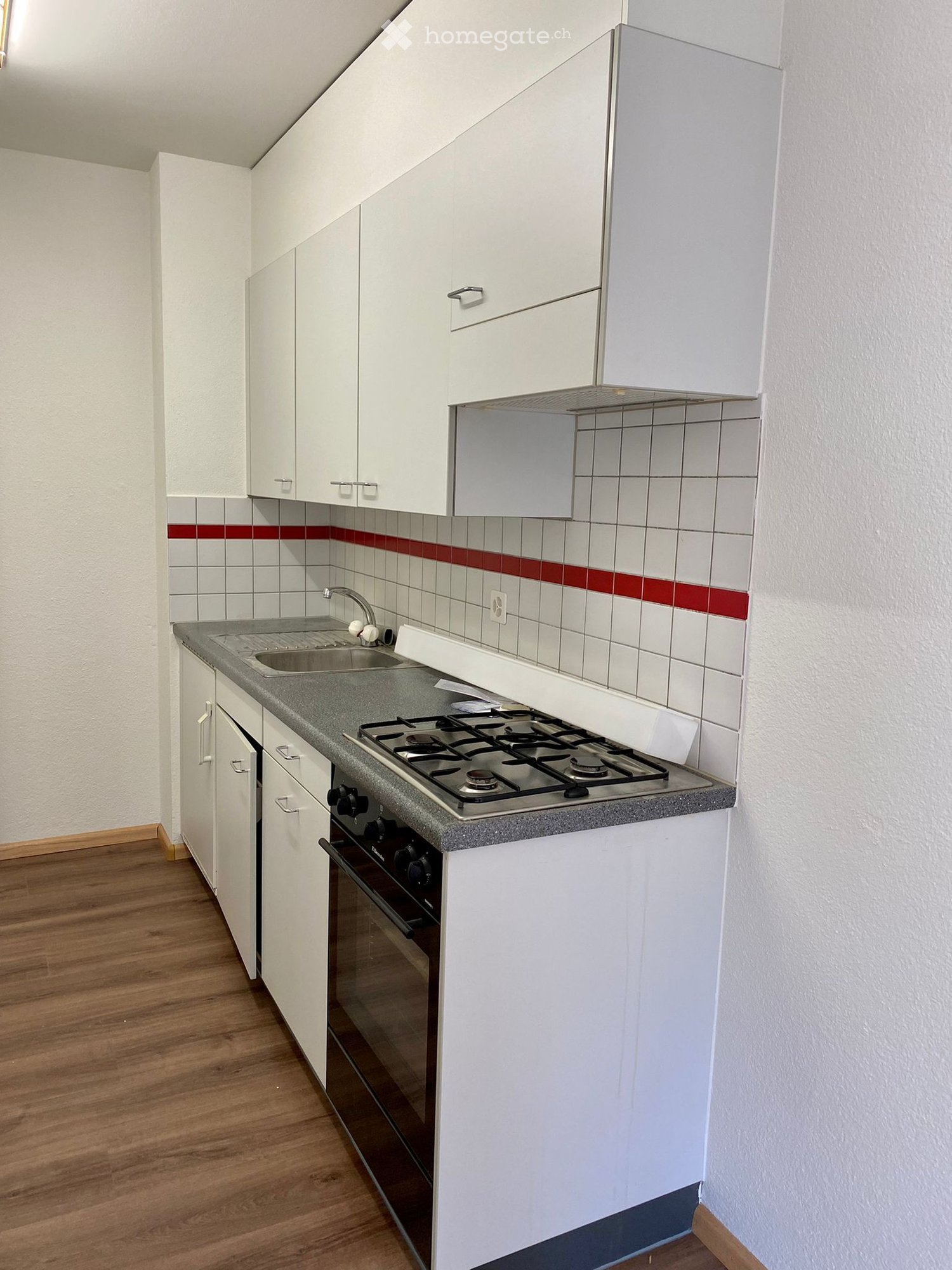 Modern kitchen with white cabinets, a gas stove, sink, and oven. The walls are tiled with a red accent stripe, and the floor is wooden.