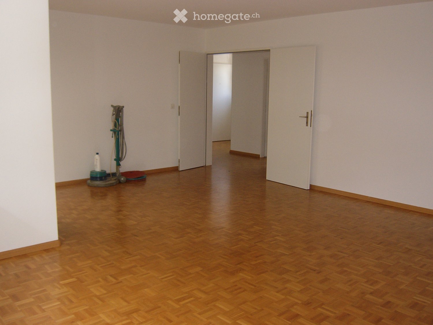 Empty room with wooden flooring and white walls, featuring a cleaning machine in the corner and an open doorway leading to another room.