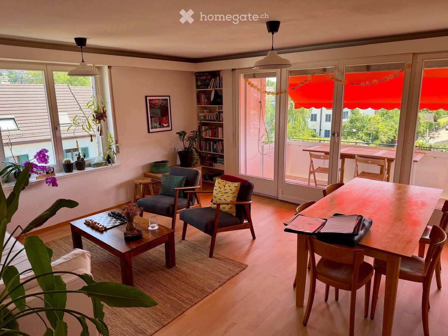 Bright living room with wooden flooring, large windows, and a balcony. Features a sofa, chairs, coffee table, dining table, bookshelf, and potted plants.