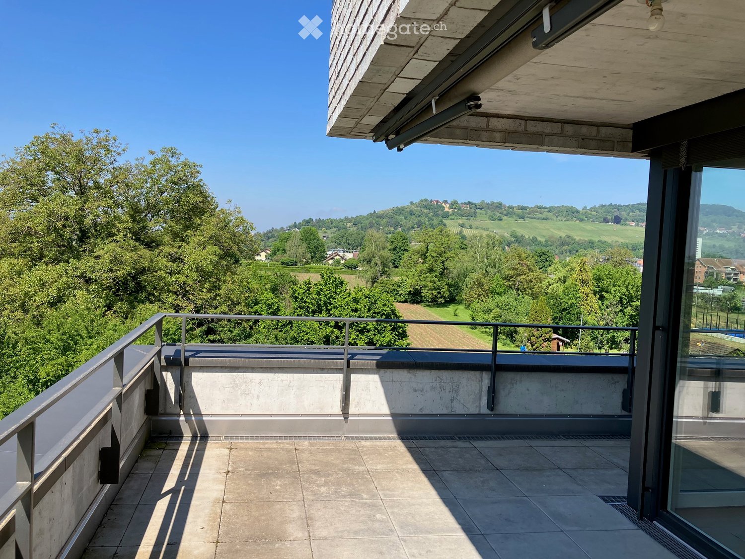 Modern balcony with concrete floor, metal railing, and expansive view of green landscape and vineyards.