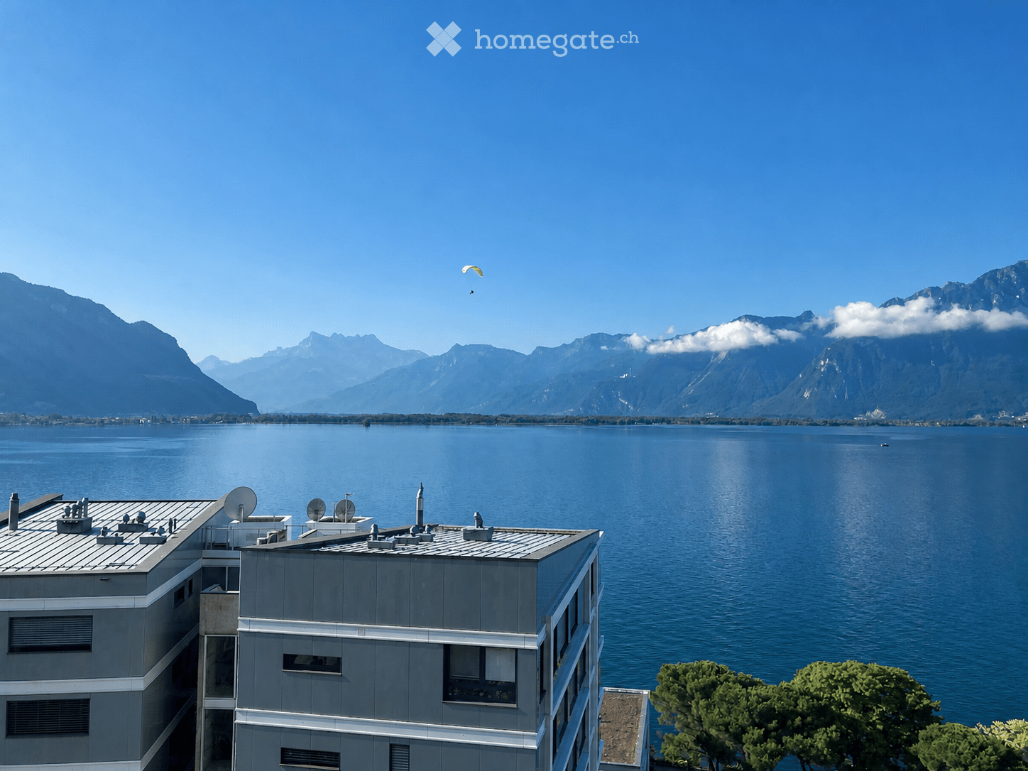 Modern multi-story building with grey facade and white trim, overlooking a large lake and mountain range under a clear blue sky.