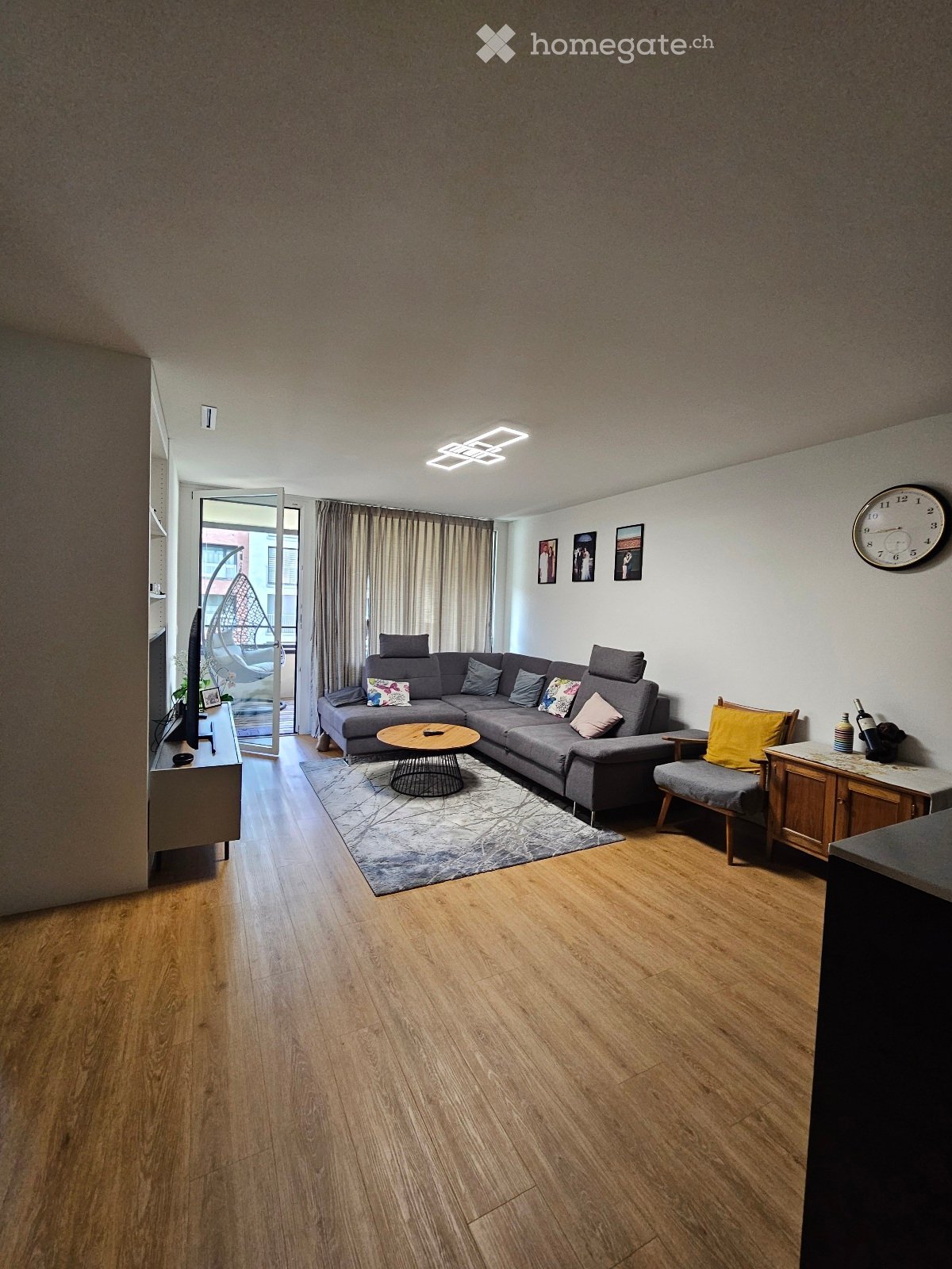 Modern living room with wooden flooring, grey sectional sofa, coffee table, side chair, and a large window with curtains leading to a balcony.