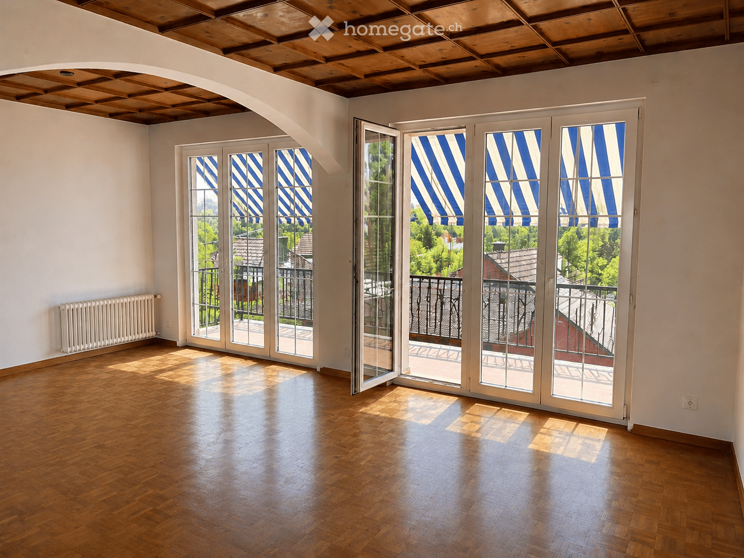 Empty room with wooden flooring, white walls, large glass doors with blue and white striped blinds leading to a balcony, and a radiator on the left wall.