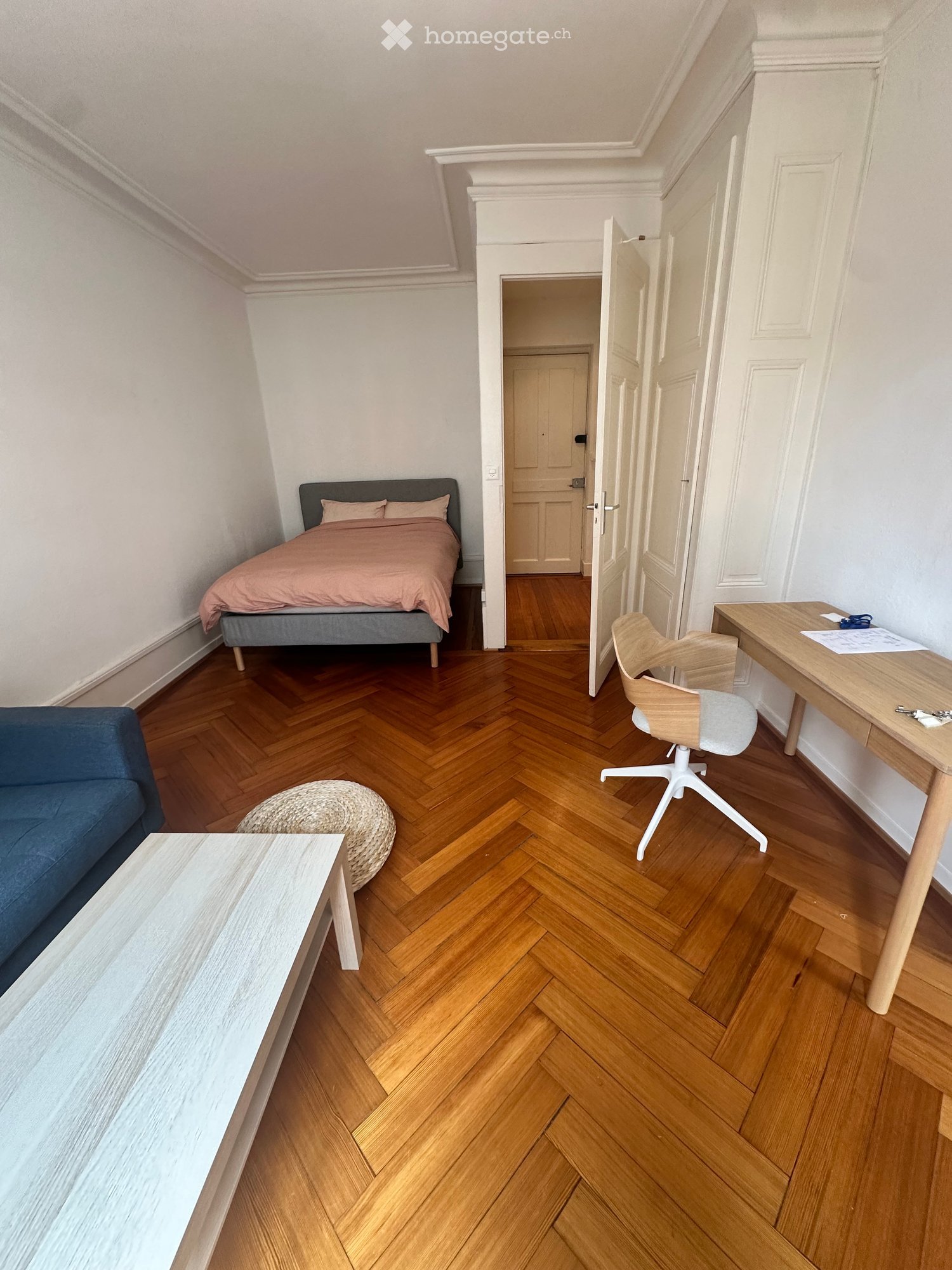 Cozy, well-furnished room with a bed, sofa, chair, table, and a round wicker stool. The room features wooden herringbone flooring and white walls.