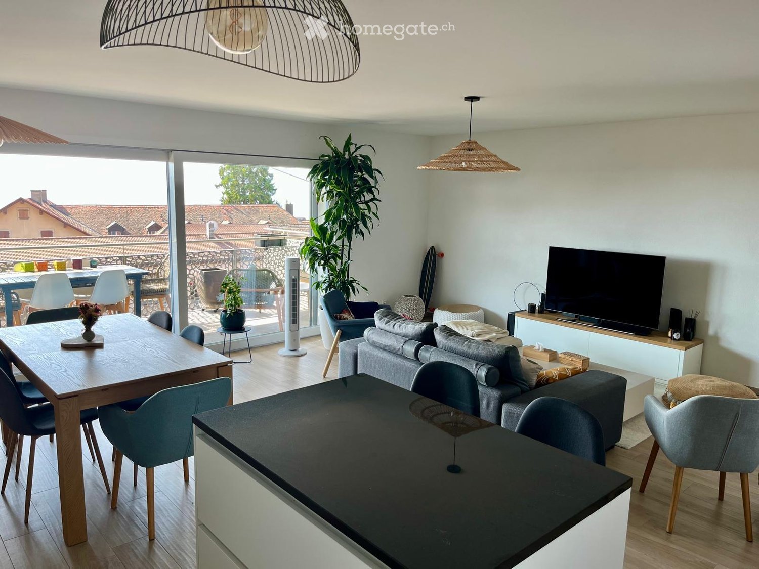 Modern living and dining area with a grey sofa, coffee table, dining table with chairs, TV, and a large plant near the balcony.