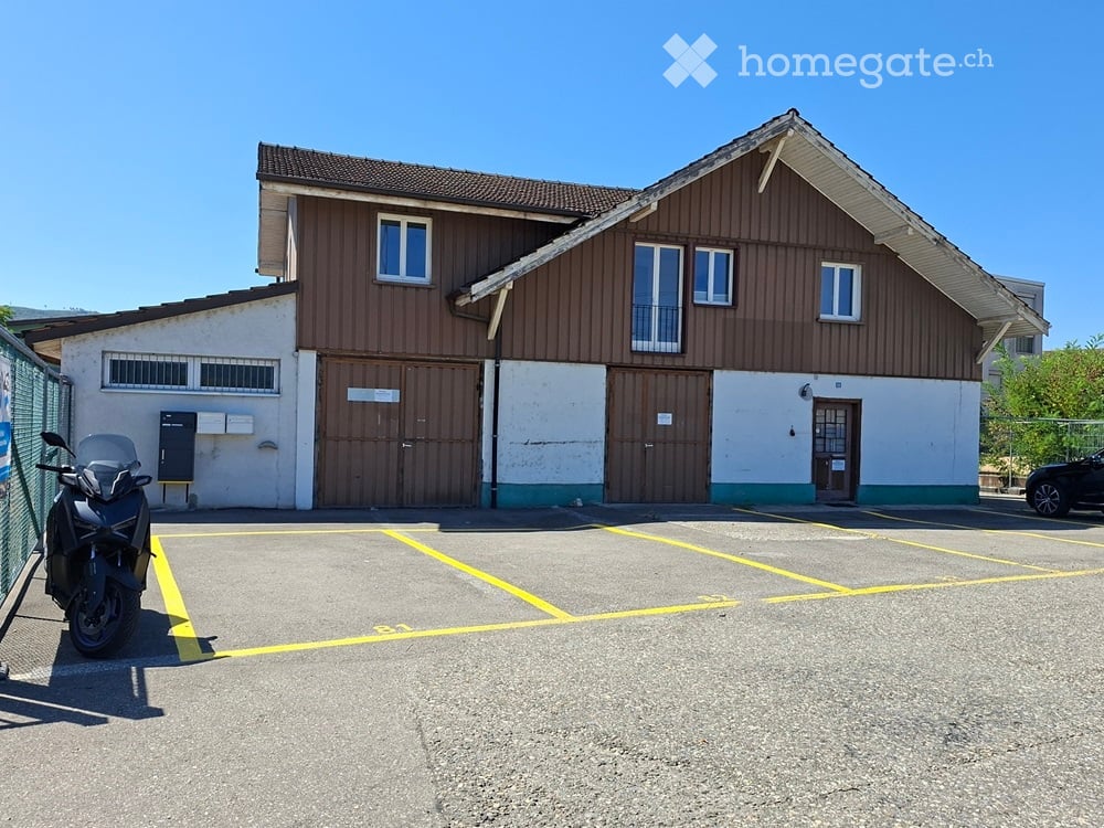 Two-story building with a garage, multiple windows, and a parking area with marked spaces.