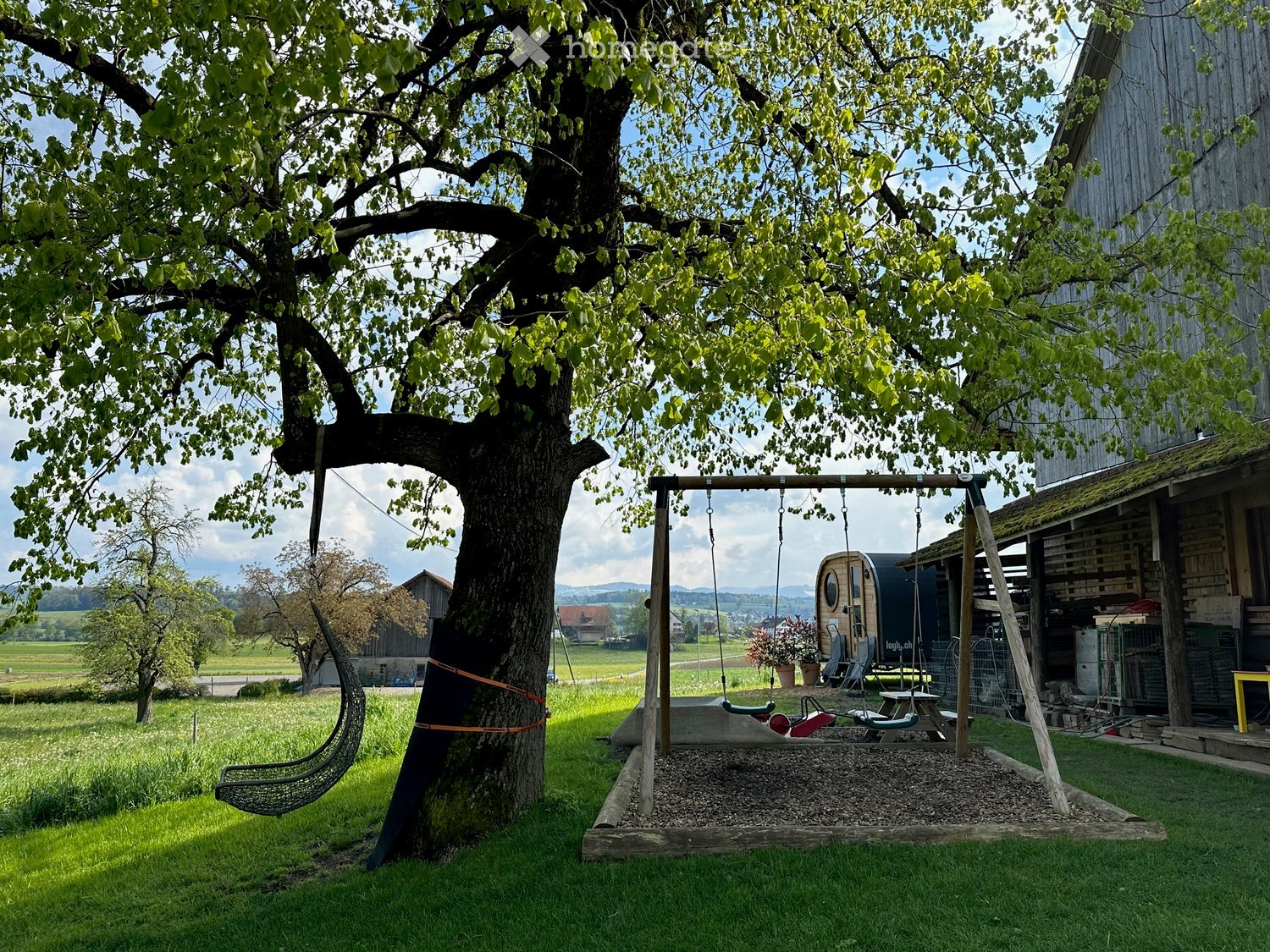 A serene garden area with a large tree, a swing attached to the tree, a wooden play structure with swings and a slide, and a small wooden cabin in the background.
