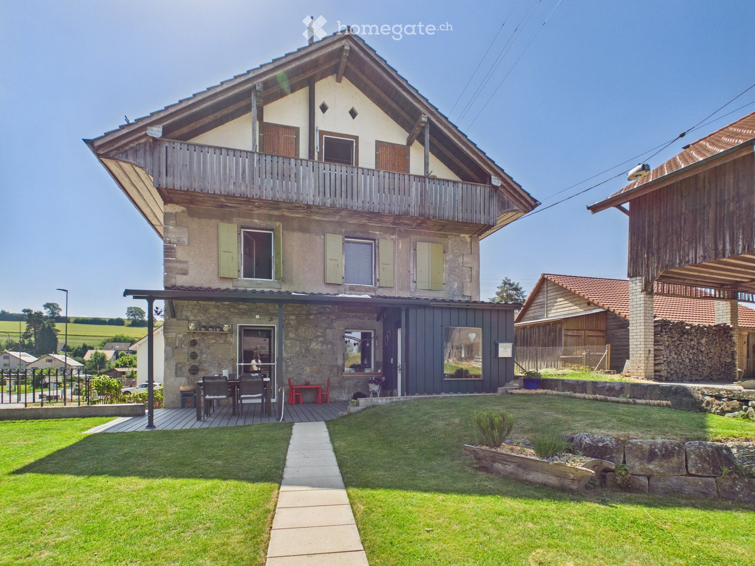 Two-story house with a balcony, stone and wood exterior, surrounded by a well-maintained lawn and garden area.