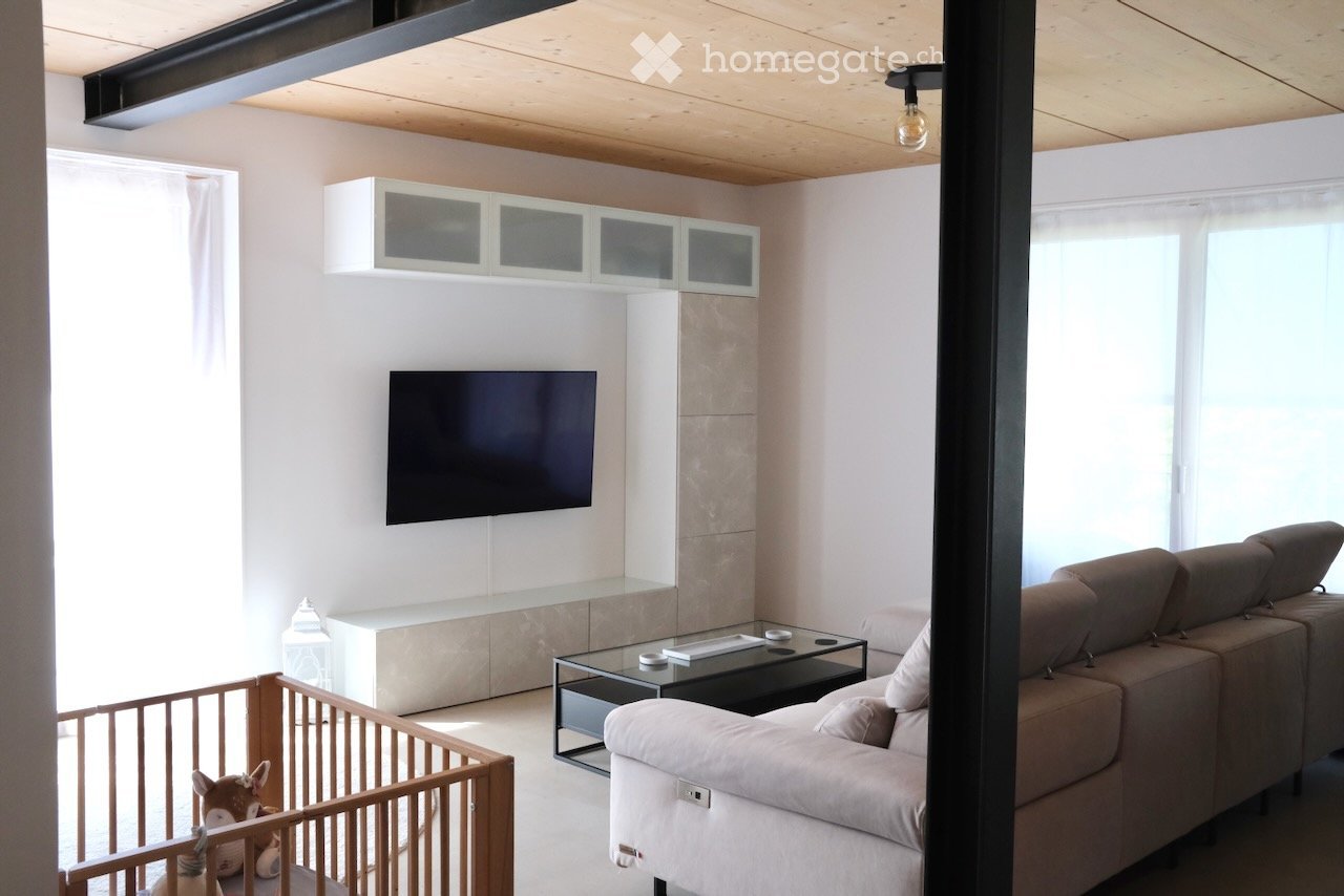 Modern living room with a grey sectional sofa, a glass coffee table, a flat-screen TV mounted on the wall, built-in white cabinets, and a small wooden playpen with a stuffed animal inside.