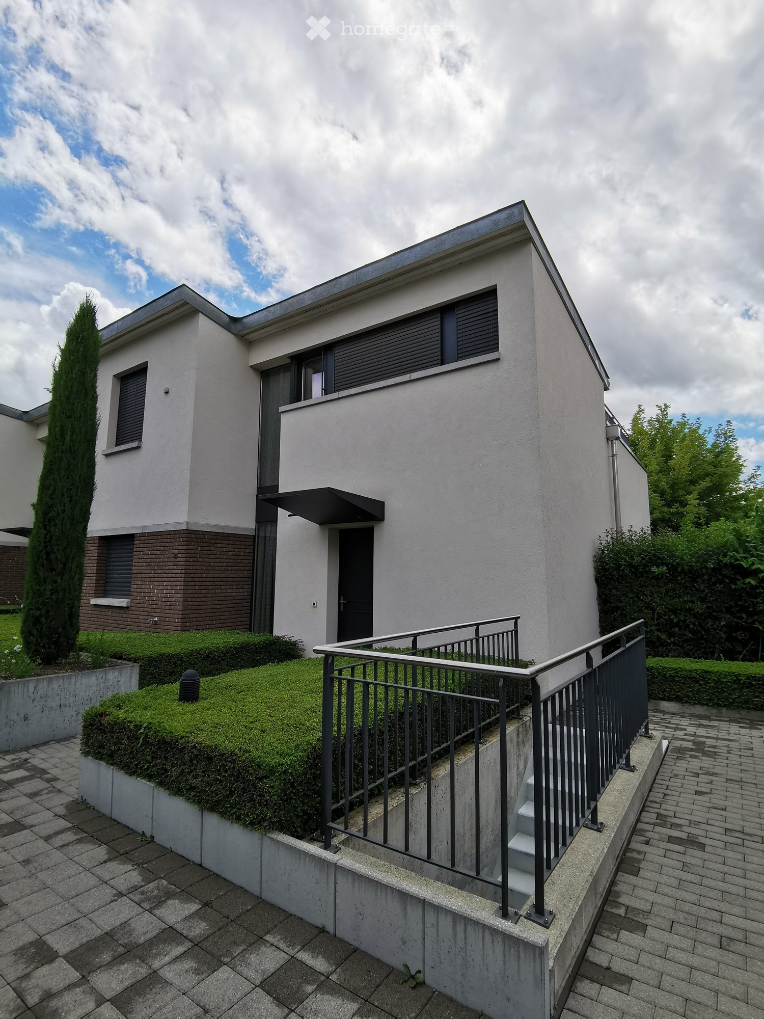 Modern two-story house with white facade, dark windows, raised ground floor, and a small staircase leading to the entrance.