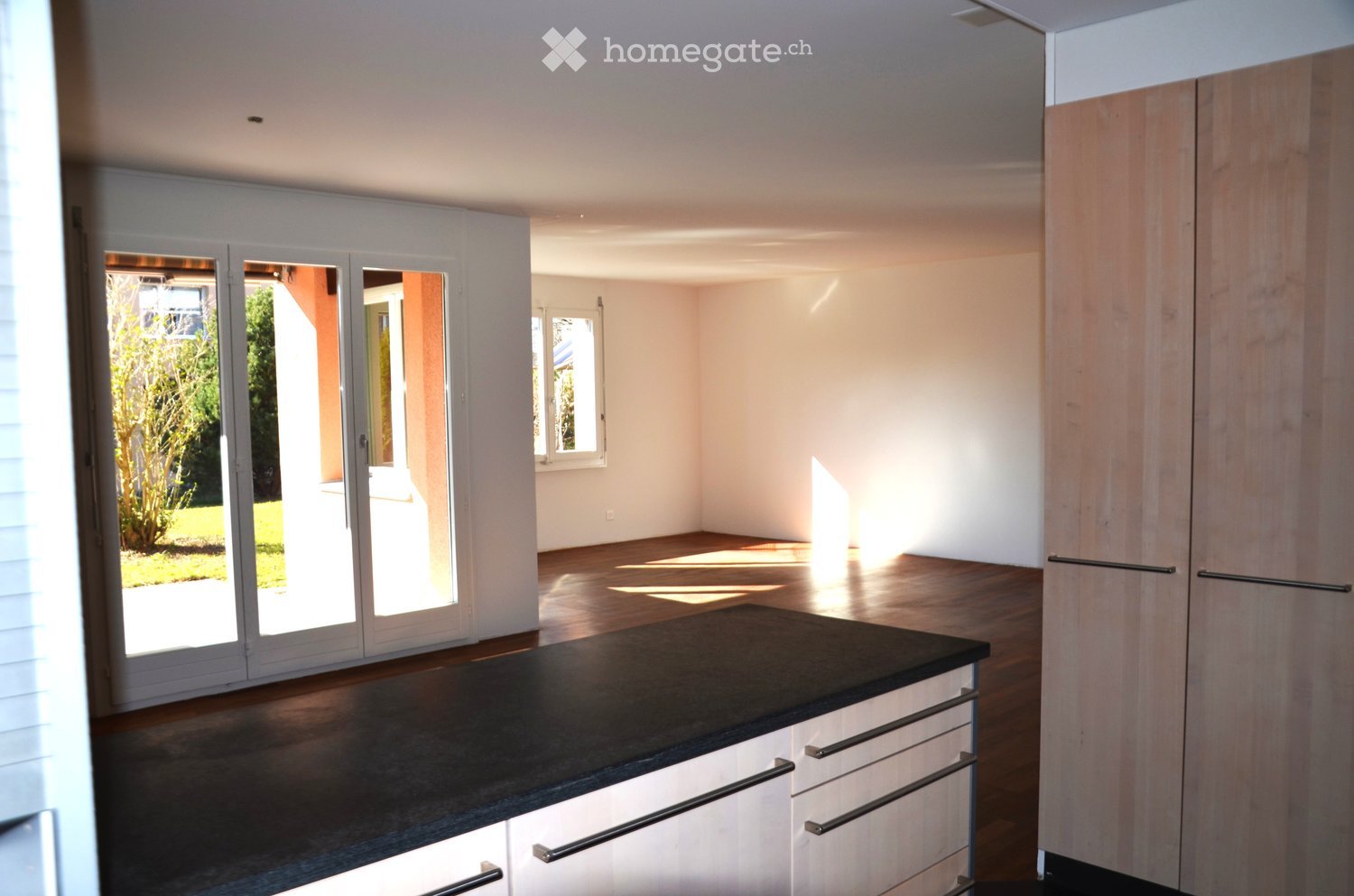 Empty room with wooden flooring, white walls, large sliding glass doors leading to a garden, and built-in white cabinetry with black countertops.