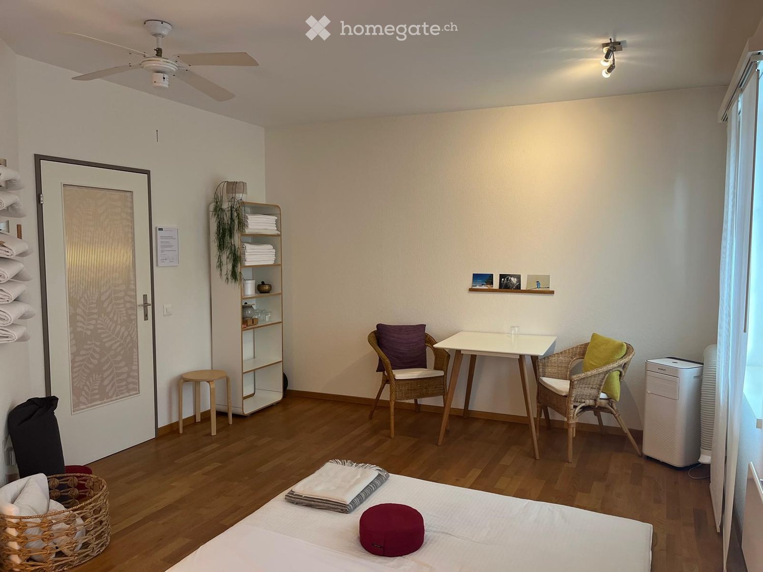 Cozy bedroom with wooden flooring, a bed with pillows and blankets, a small table with two chairs, a shelf with folded towels, a basket, and a ceiling fan.
